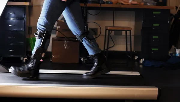A person wearing sturdy boots and jeans walks on a treadmill wearing a metal brace from the ankle to just below the knee.