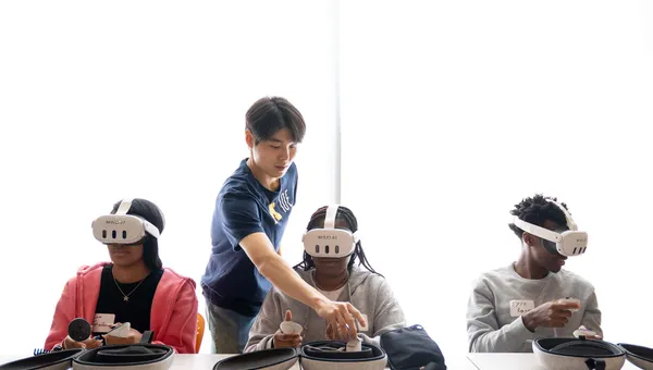 Three high school students sit at a table using VR headsets; one U-M IOE student assists one of the students.