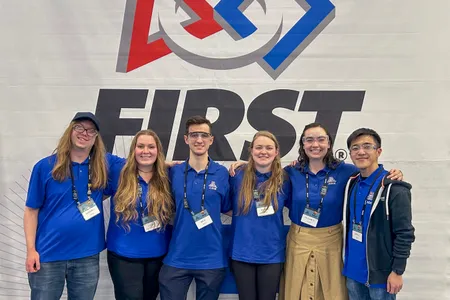 Volunteers pose in front of a FIRST Robotics banner at a competition.