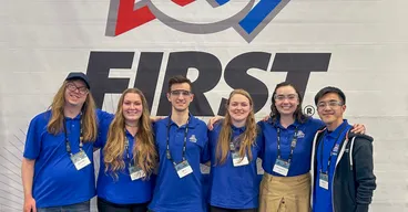 Volunteers pose in front of a FIRST Robotics banner at a competition.