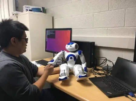 A researcher sits at a desk in front of two computers while extending his hand to the hand of a small humanoid robot, about two feet tall, that sits on the desk.
