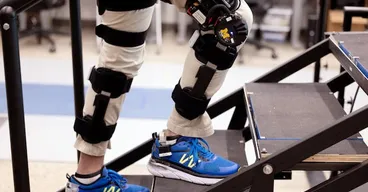 A researcher wearing motorized knee exoskeletons on both legs ascends a flight of stairs.