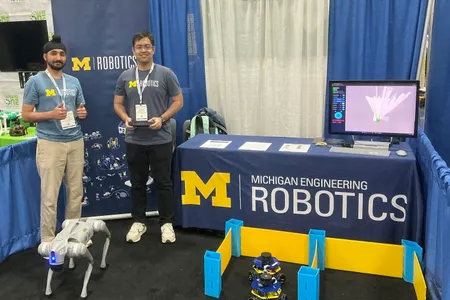 Trushant Adeshara and Manveer Singh stand next to a booth table for Michigan Robotics while demonstrating a small mobile robot and a quadruped robot at a conference.
