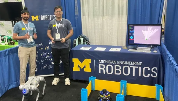 Trushant Adeshara and Manveer Singh stand next to a booth table for Michigan Robotics while demonstrating a small mobile robot and a quadruped robot at a conference.