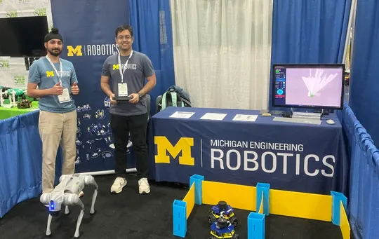 Trushant Adeshara and Manveer Singh stand next to a booth table for Michigan Robotics while demonstrating a small mobile robot and a quadruped robot at a conference.