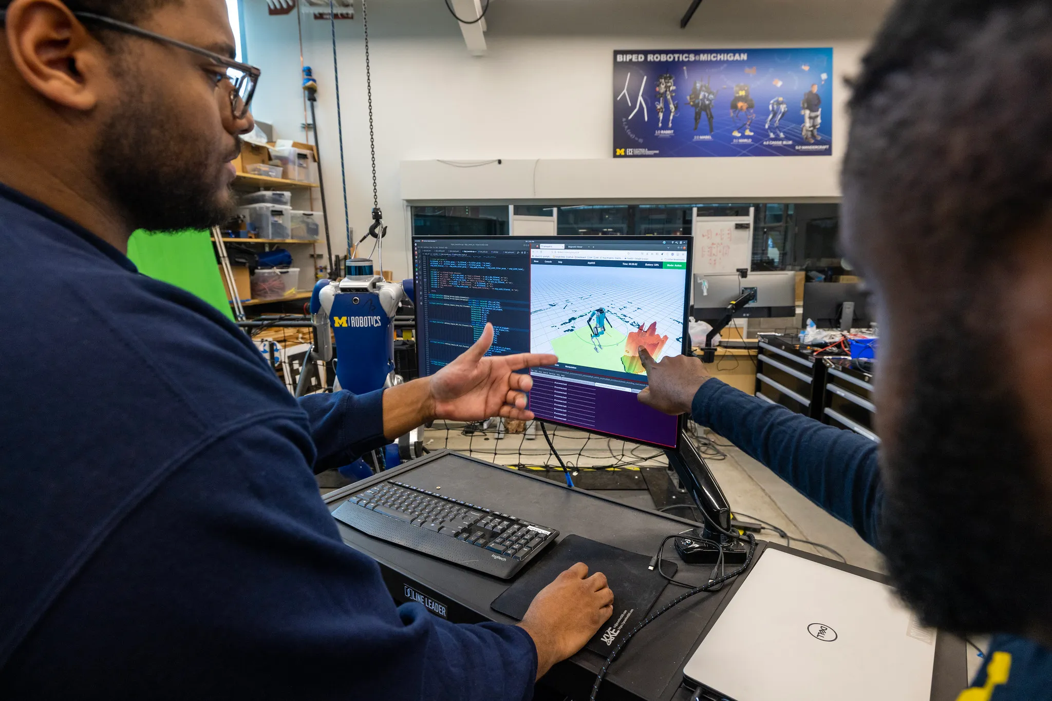 PhD students observe a simulation of a bipedal robot before testing it out on the real robot in the background.