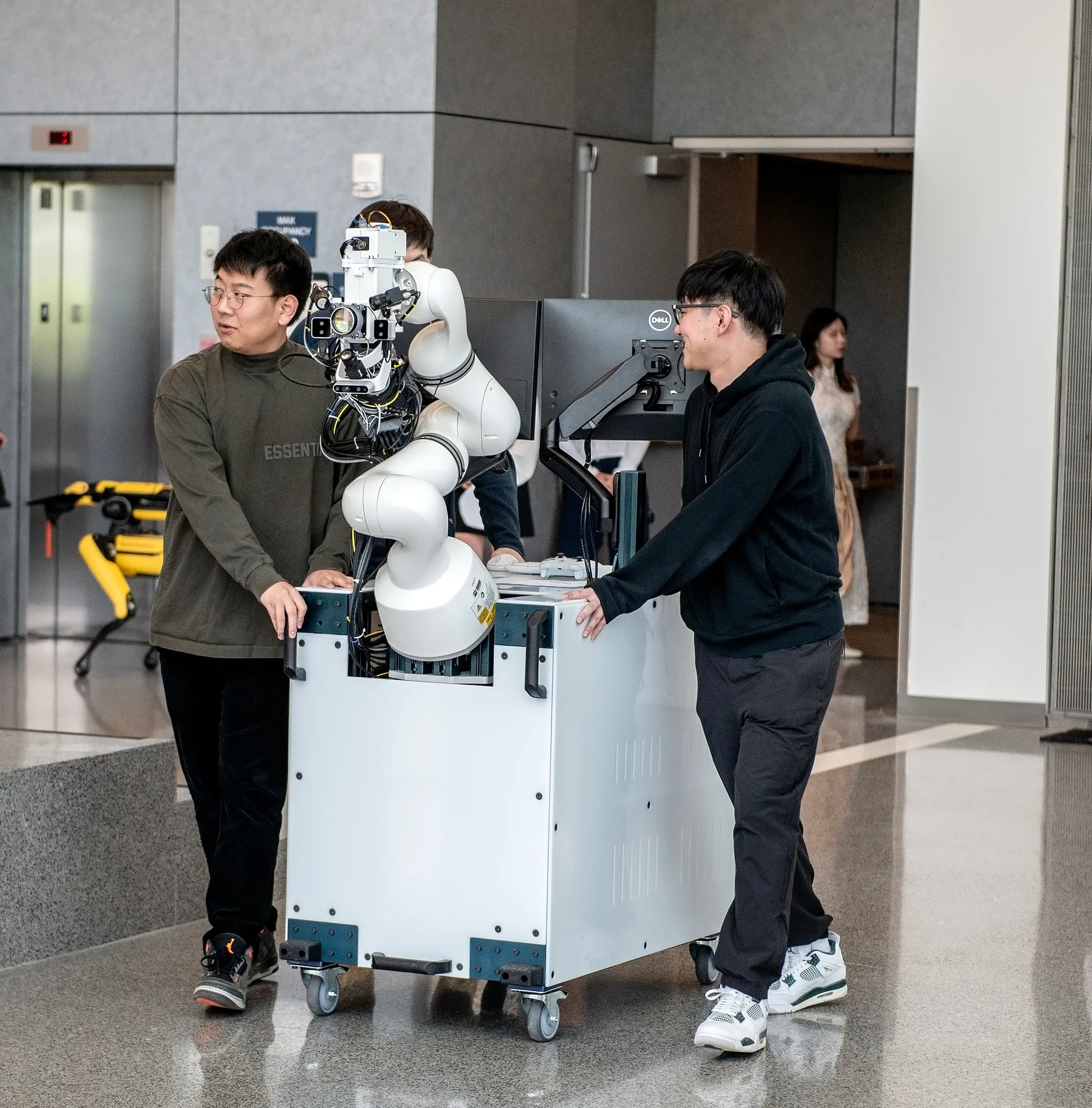 Two researchers wheel the robotic cart system made up of a robotic arm, eye image scanner, computer and monitor, through the Robotics Building atrium.