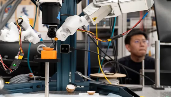 A graduate student in the background operates two robotic microsurgery arms in the foreground that are operating on the thin skin of an egg.
