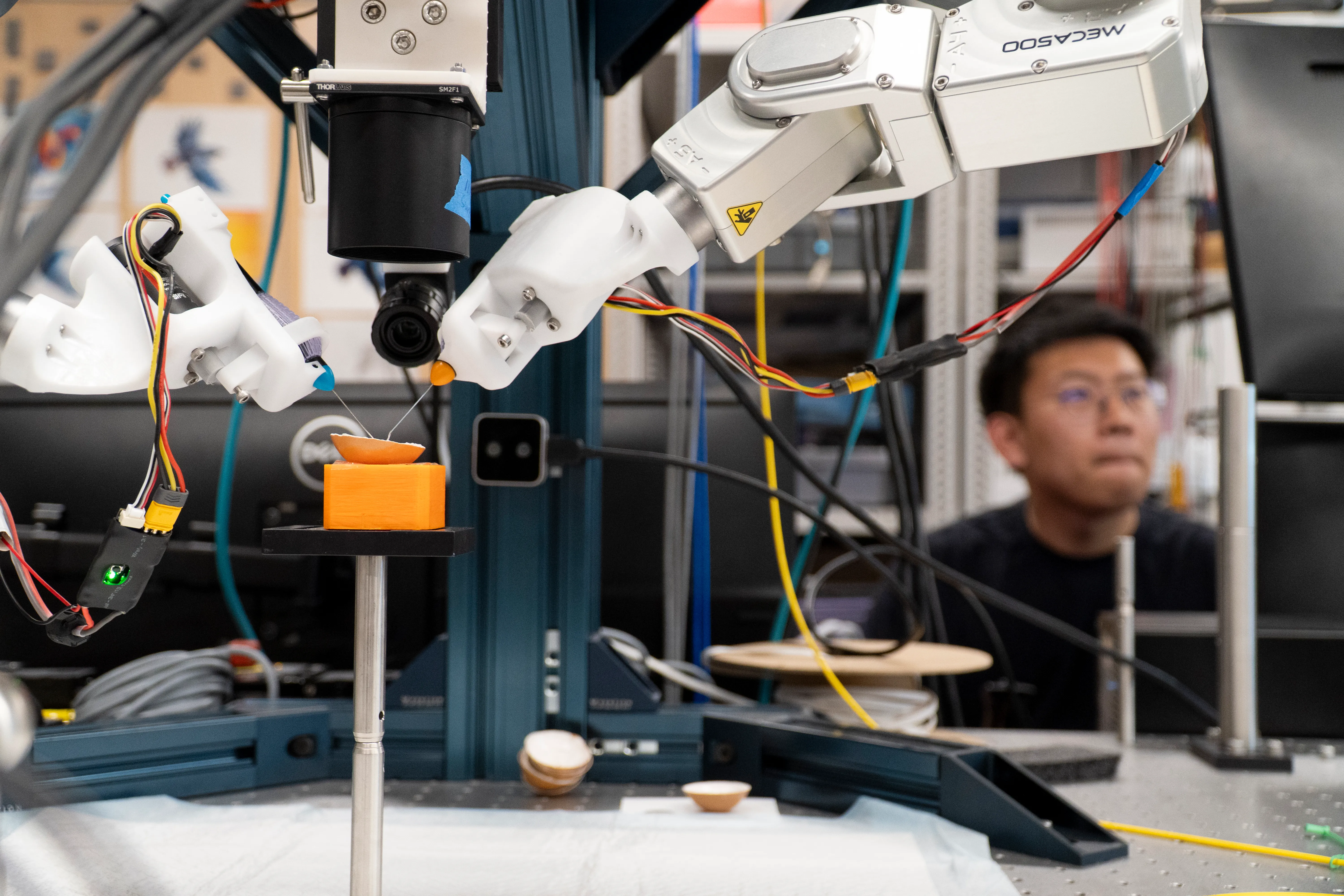 A graduate student in the background operates two robotic microsurgery arms in the foreground that are operating on the thin skin of an egg.