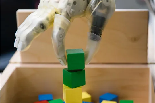Close up of advanced prosthetic hand with a green block grasped between the thumb and index finger, positioning the block at the top of a stack.