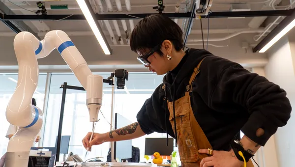 An artist paints opposite of a gleaming white robot arm, also painting on the same canvas in a research lab.