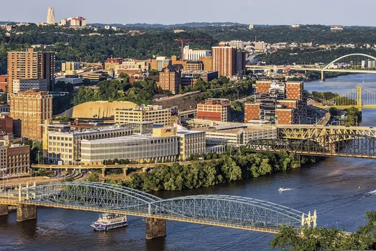 Bridges cross the Monongahela River in Pittsburgh in evening light, with a riverboat passing below and the Cathedral of Learning on the horizon.