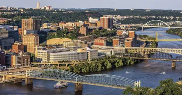 Bridges cross the Monongahela River in Pittsburgh in evening light, with a riverboat passing below and the Cathedral of Learning on the horizon.