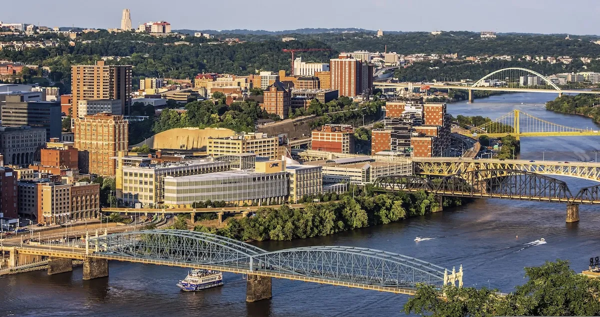 Bridges cross the Monongahela River in Pittsburgh in evening light, with a riverboat passing below and the Cathedral of Learning on the horizon.