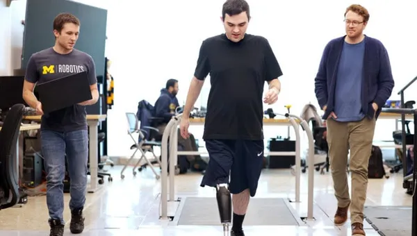 Three men walk through a lab with a treadmill built into the floor behind them. In the center, Seelhoff wears a prosthetic leg that stores its electronics inside a slim calf-shaped casing. On the left, Kevin carries an open laptop computer.