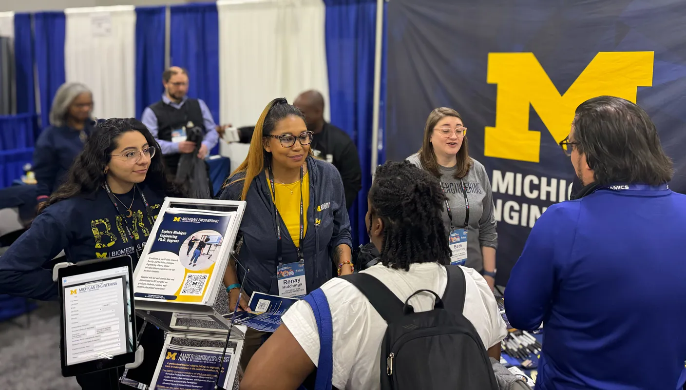 Renay Hutchings stands with other Michigan Engineering representatives at a NSBE conference booth talking to visitors.