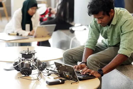 A student programs a wheeled robot on a laptop in a robotics classroom