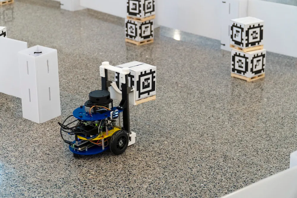 An MBot educational robot with two wheels and a forklift carries a small box on a pallet